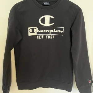 Svart Champion sweatshirt New York - Svart sweatshirt från Champion med stor vit logga och texten 'New York' på bröstet. Tröjan har rund hals, långa ärmar och en klassisk passform. Mjuk bomullskvalitet och diskret Champion-märke på ärmen. Barnstorlek!