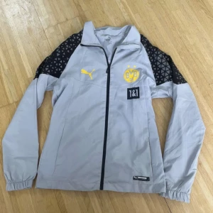 Grå vindjacka Puma BVB - Grå vindjacka från Puma med Borussia Dortmund-logga och gula detaljer. Svarta partier med geometriskt mönster på axlarna, hel dragkedja framtill och elastiska muddar. Perfekt för dig som gillar fotboll och sportig stil.