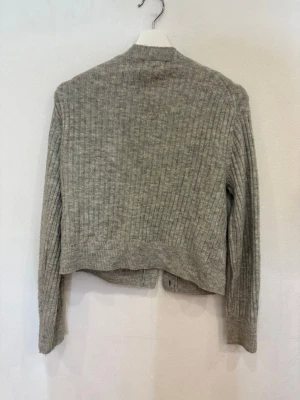 Grå ribbad kofta från H&M - Snygg grå kofta från H&M med ribbad struktur och tre stora knappar framtill. Koftan har v-ringad hals och lång ärm, perfekt för lager-på-lager. Mjuk och skön, tillverkad i stickad material som håller dig varm under kyliga dagar.