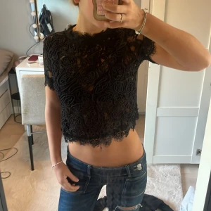Svart spetsig croppad blus - Säljer en svart croppad blus med korta ärmar och vackert spetsmönster. Toppen har genombruten design med broderade detaljer och en rå, vågig kant nertill. Perfekt för dig som vill ha en edgy och trendig look.❤️