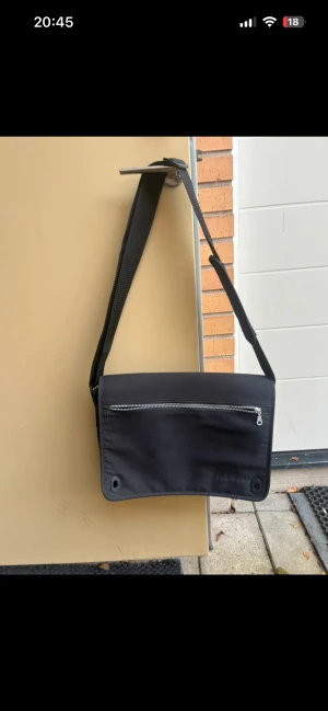 Svart axelväska med dragkedja - Snygg svart axelväska från IKEA med justerbar rem och en stor ficka med silverfärgad dragkedja på framsidan. Väskan har en stilren, rektangulär form och är tillverkad i slitstark syntet. Perfekt för att bära med sig det viktigaste.