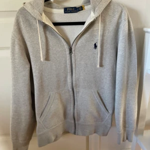 Grå Polo Ralph Lauren zip hoodie - Säljer en stilren grå hoodie från Polo Ralph Lauren, med dragkedja och klassisk logga på bröstet. Tröjan har långa ärmar och en praktisk huva med snören. Perfekt för en avslappnad look. Väldigt populär på marknaden, säljs inte längre i de flesta butikerna, alltså väldigt sällsynt. Storleken är S, men kan även passa M. Endast använd ett fåtal gånger då den var för liten, alltså nyskick. 