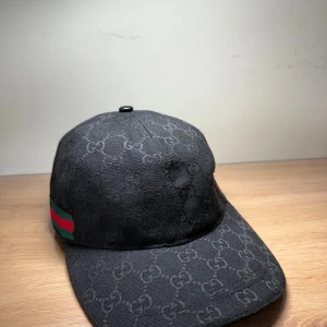 Svart Gucci keps med GG-mönster - Snygg svart keps från Gucci med klassiskt GG-monogram över hela ytan och grönt/rött band på sidan. Kepsen har böjd skärm och justerbar rem baktill. Tillverkad i bomull och polyester, perfekt för dig som gillar exklusiva streetwear-vibes.