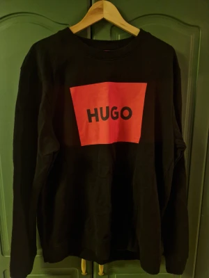 Svart sweatshirt från Hugo med röd logga XL - Svart sweatshirt från Hugo i XL med stor röd logga framtill. Tröjan har rund halsringning, långa ärmar och en relaxed passform. 