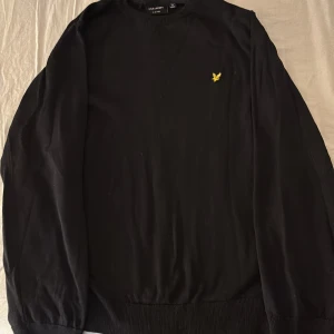 Svart stickad tröja Lyle & Scott - Svart stickad tröja från Lyle & Scott med klassisk gul broderad logga på bröstet. Rund hals och ribbade muddar vid ärmslut och nederkant. Perfekt för dig som gillar stilrena och enkla plagg med en snygg detalj.