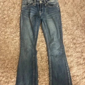 Blå bootcut jeans från Fire & Rain - Snygga blå jeans från Fire & Rain med bootcut-modell och markerade kontrastsömmar. Jeansen har klassiska fem fickor, dekorativa knappar på bakfickorna och en lätt tvättad look. Perfekta för dig som gillar en tidlös denimstil.