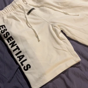 Vita Essentials shorts med snörning - Säljer ett par vita shorts från Essentials med svart logotyp längs benet. De har elastisk midja med snörning och en bakficka. Mjukt material som känns skönt mot huden, perfekt för chill dagar. Snygg och enkel design som passar till streetwear.