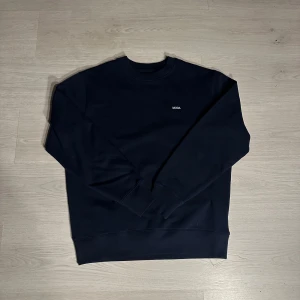 Mörkblå sweatshirt från MODA - Snygg mörkblå sweatshirt från MODA med enkel design och liten vit logga på bröstet. Tröjan har rund hals, långa ärmar och ribbade muddar vid ärmslut och nederkant. Perfekt för dig som gillar stilrena och bekväma plagg.