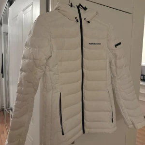 Vit pufferjacka från Peak Performance - Säljer en snygg vit pufferjacka från Peak Performance med huva och svarta detaljer. Jackan har dragkedja framtill, två fickor med dragkedja och diskret logga på bröstet. Perfekt för kalla dagar när du vill hålla stilen.