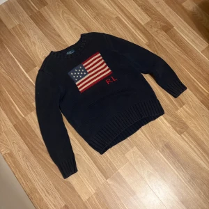 Mörkblå stickad tröja Polo Ralph Lauren - Snygg mörkblå stickad tröja från Polo Ralph Lauren med amerikansk flagga och RL-broderi på bröstet. Tröjan är i 100% bomull, har rund hals och långa ärmar med ribbade muddar. Perfekt för dig som gillar klassisk och cool stil.