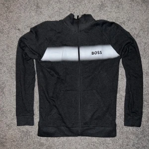Svart zip hoodie från BOSS - Säljer en svart hoodie från BOSS med hel dragkedja och vit panel över bröstet där loggan är tryckt. Tröjan har långa ärmar, ribbade muddar och fickor framtill. Perfekt för en chill och stilren look.