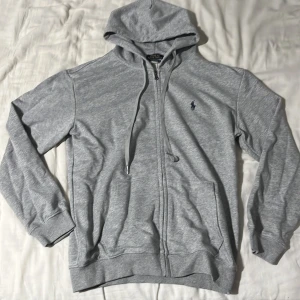 Grå hoodie från Polo Ralph Lauren - Replika!! Helt ny passar Storlek S. Snygg grå hoodie från Polo Ralph Lauren med dragkedja och huva. Klassisk broderad logga i mörkblått på bröstet, ribbade muddar och fickor framtill. Perfekt för chill dagar och enkel att matcha med jeans eller joggers.
