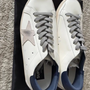 Golden Goose Superstar sneakers vit/grå - Golden Goose Superstar sneakers i vitt skinn med grå mockastjärna på sidan, grå snörning och mörkblå häl. Klassisk rund tå och platt sula med vintage-look. Snygg italiensk design med GGDB/SSTAR-tryck på sidan.
