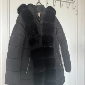 Svart pufferjacka med pälsdetaljer - Snygg svart pufferjacka med fluffiga pälsdetaljer längs huvan och framtill. Jackan har dragkedja, två fickor med dragkedja och en längre passform som håller dig varm. Perfekt för kalla dagar och ger en lyxig vibe till din outfit.