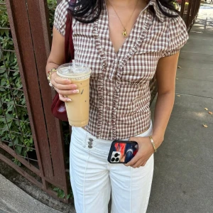 Brun- och vitrutigt blus med volang - Supersöt brun- och vitrutigt blus med korta puffärmar, markerad midja och volangdetaljer längs knäppningen. Skjortan har en djup v-ringning och klassisk krage. Perfekt för dig som gillar vintage vibes och en feminin look.