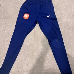 Blå Nike träningsbyxor  - Säljer ett par blå Nike träningsbyxor i slim fit-modell med Dri-FIT-material. Byxorna har resår i midjan, snörning och ett orange lejonmärke på ena benet. Perfekta för dig som gillar sportig stil och vill ha bekväma byxor till träning.