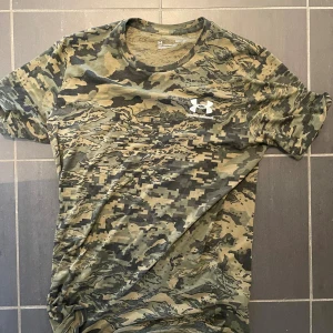 Camouflage t-shirt från Under Armour - Snygg t-shirt från Under Armour med digitalt camouflage-mönster i gröna och beige toner. Klassisk rund hals och korta ärmar. Perfekt för dig som gillar sportig stil och vill sticka ut med ett coolt mönster.