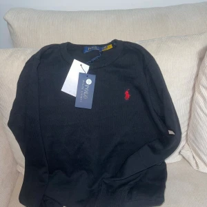 Polo Ralph Lauren stickad tröja - Klassisk stickad tröja från Polo Ralph Lauren med rund hals och broderad logga på bröstet. Finns i svart med röd logga och grå med marinblå logga. Tröjan har ribbade muddar och är perfekt för lager-på-lager. Snygg och tidlös design.