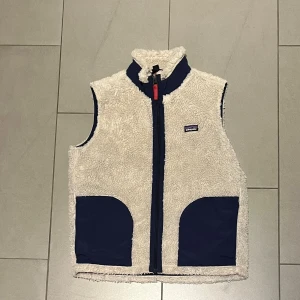 Vit fleeceväst från Patagonia - Säljer en vit fleeceväst från Patagonia med marinblå detaljer och fickor. Västen har hög krage, hel dragkedja och mjukt, fluffigt material på utsidan. Insidan är fodrad med marinblå polyester. Perfekt för lager-på-lager och chill stil.