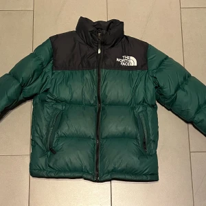 Grön pufferjacka från The North Face - Säljer en grön och svart pufferjacka från The North Face med hög krage och två sidofickor med dragkedja. Jackan har en tydlig logga på bröstet och axeln, quiltad design och hel dragkedja framtill. Perfekt för kalla dagar och har en riktigt chill vibe.