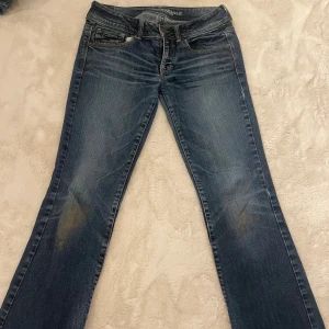 Blå bootcut jeans från American Eagle - Snygga blå jeans från American Eagle med klassisk bootcut passform. Jeansen har fem fickor, slitningar och kontrastsömmar. Materialet är jeans med en skön stretch och de har låg midja. Bakfickorna har broderade detaljer. Har ett litet litet hål vid inne låret. Köpte för 800.