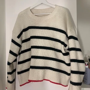 Randig stickad tröja i vitt och svart - Säljer en stickad tröja med svarta ränder på vit botten och en röd detalj längst ner. 