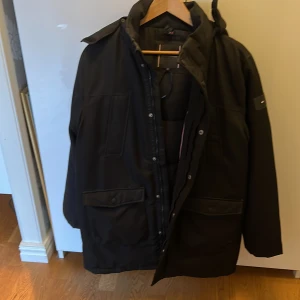 Svart parkas från Tommy Hilfiger - Svart parkas från Tommy Hilfiger med pufferfoder och huva med snygg pälsdetalj. Jackan har stora fickor med lock och knappar, dragkedja och tryckknappar framtill. Perfekt för kalla dagar med sin långa modell och varma material.