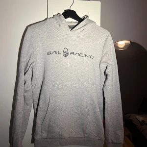 Grå hoodie från Sail Racing - Snygg grå hoodie från Sail Racing med svart logotyp tryckt på bröstet. Tröjan har huva, lång ärm och en stor magficka framtill. Mjuk och skön bomullsmix som passar perfekt när du vill chilla eller hänga med kompisar.
