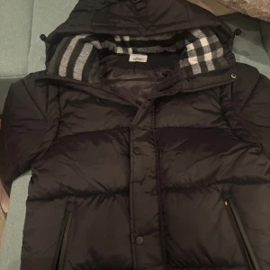 Svart Burberry jacka  - Svart pufferjacka från Burberry med avtagbara ärmar så den kan användas som väst. Jackan har huva med grått och vitt rutigt foder, dragkedja och tryckknappar framtill samt två sidofickor med dragkedja. Stor Burberry London England-logga på bröstet.