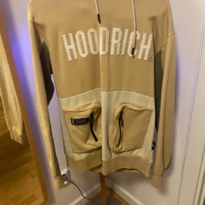 Beige hoodie från Hoodrich med fickor - Säljer en beige och ljusbeige hoodie från Hoodrich i storlek M. Tröjan har stor logga framtill, två stora fickor med dragkedja och snörning i huvan. Perfekt för dig som gillar streetwear och vill ha en snygg och bekväm look.
