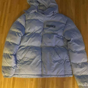 Ljusblå pufferjacka från Hoodrich - Säljer en fet ljusblå pufferjacka från Hoodrich med stor logga på ryggen och mindre logga på bröstet. Jackan har huva, två stora fickor fram och breda kanaler. Perfekt för kalla dagar när du vill se fresh ut.