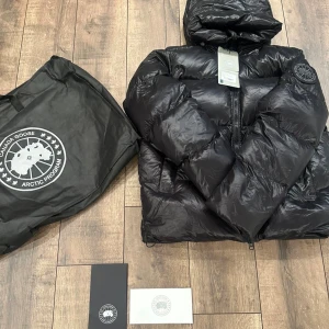 Svart pufferjacka från Canada Goose - Säljer en svart pufferjacka från Canada Goose med huva och tydlig logotyp på ärmen. Jackan har dragkedja framtill, stora fickor och justerbara remmar på insidan. Materialet är glansigt och dunfyllt, perfekt för kalla dagar. Medföljer originalpåse och märkeskort.