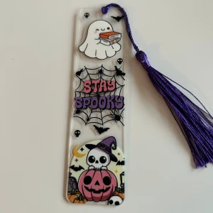 Stay Spooky (bokmärke) - Ett färgglatt bokmärke med Halloween-tema! Pryds av ett gulligt spöke med böcker, spindelnät, fladdermöss och en söt pumpa med skelett. Perfekt för dig som älskar böcker och vill ha en spooky touch i din läsning. Lila tofs för extra stil.