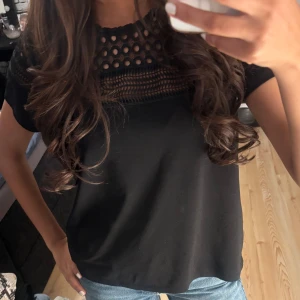 Svart blus med spetsdetaljer - Svart blus med korta ärmar och snygga spetsdetaljer upptill. Ovanför bröstet finns ett parti med hålmönstrad spets och dekorativa band. Blusen har rund hals och är lätt loose i passformen. Jättebra skick. Xs/s