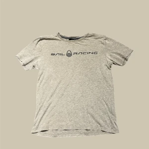 Grå t-shirt från Sail Racing - Snygg grå t-shirt från Sail Racing med svart logotyp tryckt över bröstet. Klassisk rund hals och korta ärmar. T-shirten är tillverkad i mjuk bomull som känns skön mot huden. Perfekt för dig som gillar stilren design och sportiga märken.