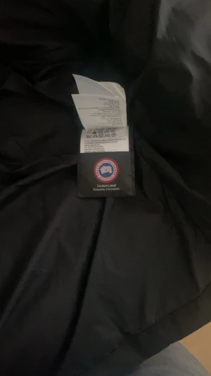 Svart dunjacka från Canada Goose - Säljer en svart dunjacka från Canada Goose med huva och klassisk logga på ärmen. Jackan har lång ärm, knappar och dragkedja framtill. Perfekt för kalla dagar med sin tjocka och puffiga design. Stilsäker och funktionell för vintern.