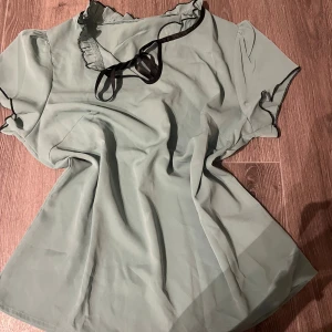 Grön blus med svarta detaljer - Snygg ljusgrön blus med svarta kanter och rosett vid halsen. Blusen har korta ärmar och volangdetaljer längs halsringningen och ärmsluten. Materialet känns lätt och luftigt, perfekt för dig som gillar en chill och feminin stil. Man kan även knyta upp och styla knytningen vid halsen hur man vill.