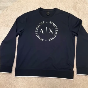 Mörkblå sweatshirt från Armani Exchange - Snygg mörkblå sweatshirt från Armani Exchange med stort vitt logotryck på bröstet. Tröjan har rund hals, långa ärmar och vita detaljer vid muddar och nederkant. Tillverkad i mjuk bomull för skön komfort. Perfekt för dig som gillar stilrena och trendiga plagg.