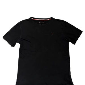 Svart t-shirt från Tommy Hilfiger - Svart t-shirt från Tommy Hilfiger med diskret logga på bröstet och en snygg rödvit randig detalj på insidan av halsen. Klassisk rund hals och korta ärmar. Perfekt för dig som gillar stilrena och enkla plagg med en touch av designer.