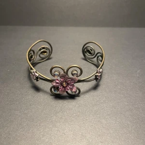 Blommigt armband med rosa detaljer - Unikt armband i antikguld-färgad metall med snirkliga former och blommor i rosa och lila. Dekorerat med glittrande stenar som ger extra bling. Blommönstret är i fokus och armbandet har en öppen design som gör det lätt att ta på och av.