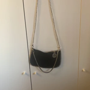 Svart axelväska med kedjedetaljer - Snygg svart axelväska med glansig yta och silverfärgade kedjor som axelrem och runt kanterna. Väskan har dragkedja upptill och en rund metallbricka som detalj. Perfekt storlek för det viktigaste och ger en cool vibe till din outfit.