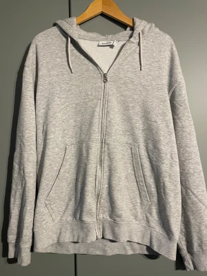 Grå hoodie med dragkedja från Weekday - Säljer en grå hoodie från Weekday i storlek M. Tröjan har huva med snörning, två fickor fram och hel dragkedja. 