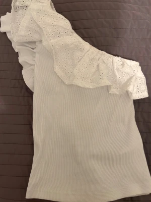 Vit oneshoulder topp med volang - Supersöt vit oneshoulder topp med bred volang i broderad spets och ribbad struktur. använd 1-2 gånger.