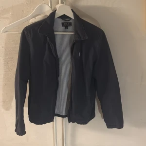 Mörkblå vindjacka från Polo Ralph Lauren -  Snygg mörkblå vindjacka från Polo Ralph Lauren med klassisk krage och dragkedja framtill. Jackan har det ikoniska broderade Polo-logot på bröstet och insidan är fodrad med ljusblå och vita ränder. Perfekt för dig som vill ha en stilren och tidlös look. Är storlek L i boys som är XS i herr storlek