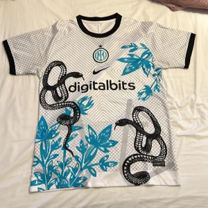 Inter Milan x Nike vit fotbollströja - Säljer en Inter Milan fotbollströja från Nike i vit med svarta och blå detaljer. Tröjan har korta ärmar, svart krage och muddar. Framsidan har grafiskt mönster med ormar och blå blommor, samt Digitalbits-logga. Materialet är lätt och andas, perfekt för match eller träning.