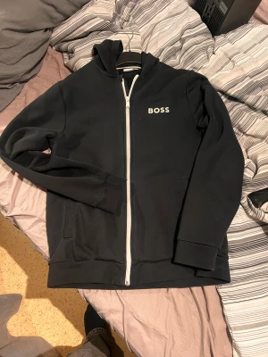 Svart hoodie från BOSS med dragkedja - Svart hoodie från BOSS i storlek M med vit dragkedja och BOSS-logga på bröstet. Tröjan har huva, långa ärmar och två fickor framtill. Passar som S eller Xs