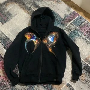 Svart hoodie med färgstark konst - Unik svart hoodie med dragkedja och huva, prydd med handmålat konstverk i blått, orange, rött och vitt på bröstet. Tröjan har ribbade muddar och en cool, konstnärlig vibe som sticker ut. Perfekt för dig som gillar streetwear med personlig touch.