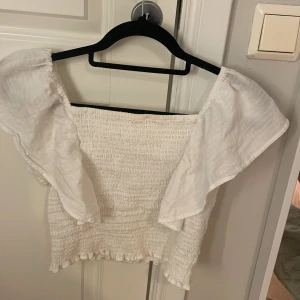 Två smockade toppar från Kappahl - Säljer två smockade toppar från Kappahl i storlek 158/164. En är vit och en är grön  båda med breda volangärmar och fyrkantig halsringning. Topparna har elastisk smock över hela kroppen och en söt volangkant nertill. Perfekta för en trendig och luftig look.