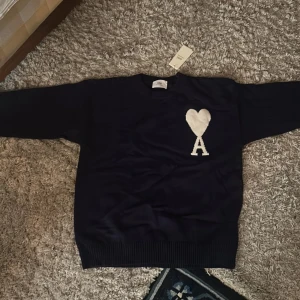 Mörkblå stickad tröja från Ami Paris - Säljer en mörkblå stickad tröja från Ami med ett vitt hjärta och bokstaven A på bröstet. Tröjan har rund hals, långa ärmar och breda ribbade muddar vid ärmslut och nederkant. Perfekt för dig som gillar stilrena detaljer och snygg design.