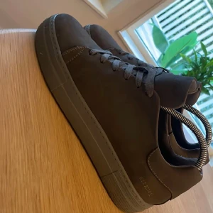 Mörkgråa sneakers från Pavement - Snygga mörkgråa sneakers från Pavement med rund tå och platt sula. Skorna är tillverkade i skinn och har klassisk snörning framtill. Perfekt för dig som gillar stilrena och tidlösa sneakers med en clean look.